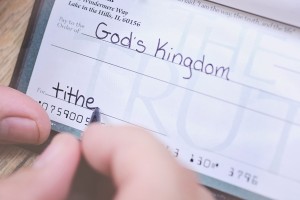 Stock photo of someone writing a check of an unspecified amount made out to "God's Kingdom" with "tithe" in the memo line