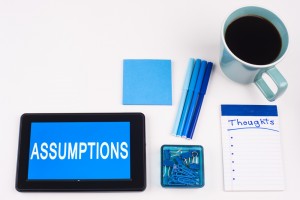 Stock photo of a tablet that has the word "ASSUMPTIONS" on it, a stack of blue post it notes, a group of blue pens, a notepad that has "THOUGHTS" written on it and a cup of coffee