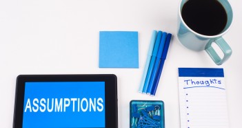 Stock photo of a tablet that has the word "ASSUMPTIONS" on it, a stack of blue post it notes, a group of blue pens, a notepad that has "THOUGHTS" written on it and a cup of coffee