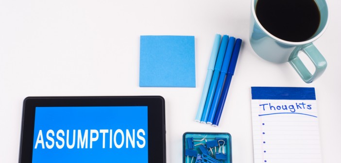 Stock photo of a tablet that has the word "ASSUMPTIONS" on it, a stack of blue post it notes, a group of blue pens, a notepad that has "THOUGHTS" written on it and a cup of coffee