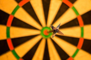 Stock photo of an empty, out-of-focus dartboard save for one, lone, in-focus dart on the bulls-eye