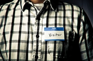Photo of an individual wearing a black-and-white checkered shirt and a nametag that reads "hello my name is VISITOR"