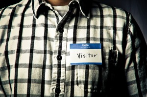 Photo of an individual wearing a black-and-white checkered shirt and a nametag that reads "hello my name is VISITOR"