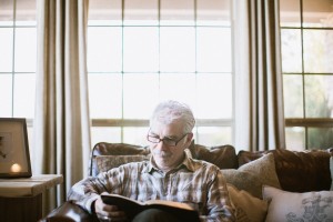 Stock photo of an older white man sitting on a sofa and reading in his living room