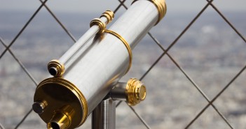 Stock photo of a silver and gold telescope looking over a city from behind a chain-fence