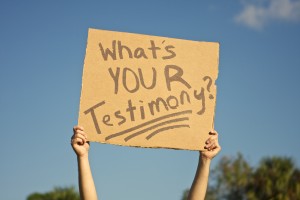 Stock art of someone holding up a cardboard sign that says "WHAT'S YOUR TESTIMONY?"