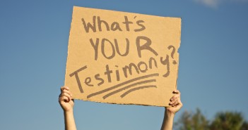 Stock art of someone holding up a cardboard sign that says "WHAT'S YOUR TESTIMONY?"