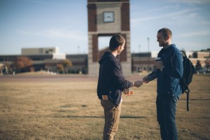 Stock photo of one young white man handing a pamphlet to another young white man in a school's quad