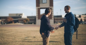 Stock photo of one young white man handing a pamphlet to another young white man in a school's quad