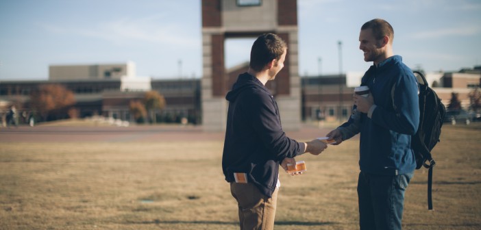 Stock photo of one young white man handing a pamphlet to another young white man in a school's quad