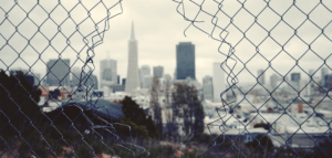 Looking through a hole in a chain link fence at a gray cityscape