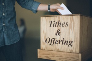 Stock photo of someone putting an envelope in a box labeled "Tithes & Offering"
