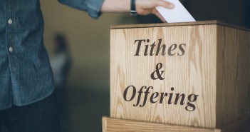 Stock photo of someone putting an envelope in a box labeled "Tithes & Offering"