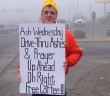 Person standing outside on the side of the road holding a sign that reads “Ash Wednesday Drive-Thru Ashes & Prayer Up Ahead on Right. Free Coffee!!”