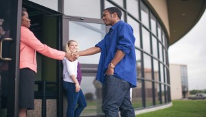 Stock photo of a middle aged African American woman in business attire shaking hands with a younger African American man dressed like a college student at the entryway of a building with a younger white woman holding the door.