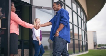 Stock photo of a middle aged African American woman in business attire shaking hands with a younger African American man dressed like a college student at the entryway of a building with a younger white woman holding the door.