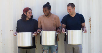 Three smiling volunteers holding large, heavy cooking pots
