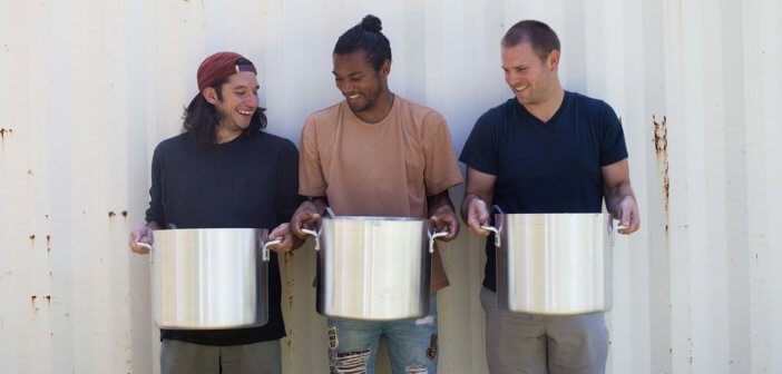 Three smiling volunteers holding large, heavy cooking pots