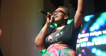 Person with a tattoo visible on their arm and wearing a t-shirt singing joyfully at church