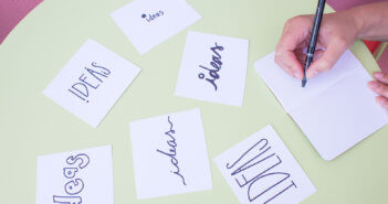 Person writing in a notebook on a table covered with index cards that say IDEAS