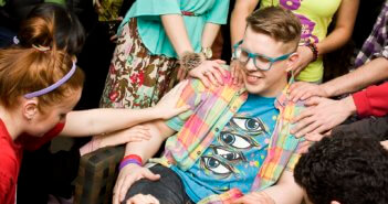 Teen sitting in chair while friends place their hands on his shoulders and arms.