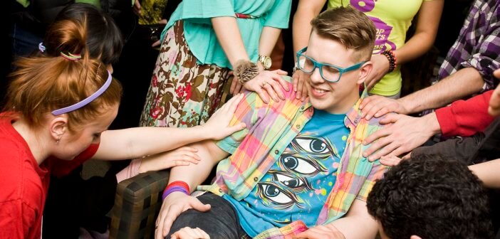 Teen sitting in chair while friends place their hands on his shoulders and arms.