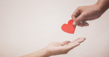 A person's hand placing a paper heart in the outstretched hand of another person