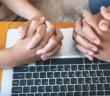 Two people with their hands clasped in prayer atop a computer keyboard