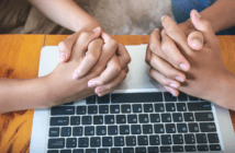 Two people with their hands clasped in prayer atop a computer keyboard