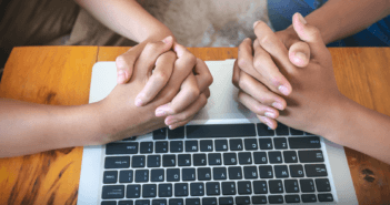 Two people with their hands clasped in prayer atop a computer keyboard