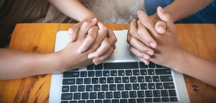 Two people with their hands clasped in prayer atop a computer keyboard