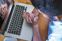 Person with clasped hands praying while engaging in an online worship service on a computer