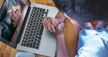 Person with clasped hands praying while engaging in an online worship service on a computer