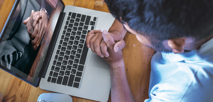 Person with clasped hands praying while engaging in an online worship service on a computer