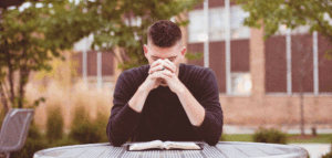 Young pastor sitting outside praying at a table with an open Bible on it