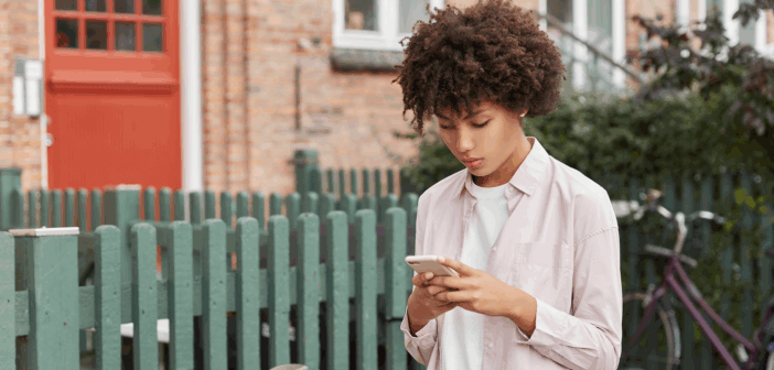 Young adult looking down at their phone while walking down the sidewalk past a fence and house