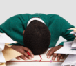 A stressed-out workaholic with their head down on their desk surrounded by piles of papers and files