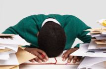 A stressed-out workaholic with their head down on their desk surrounded by piles of papers and files
