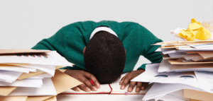 A stressed-out workaholic with their head down on their desk surrounded by piles of papers and files
