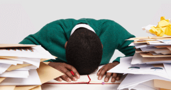 A stressed-out workaholic with their head down on their desk surrounded by piles of papers and files