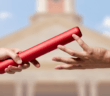 Two people's hands passing a baton from one to the other in front of a blurred image of a church