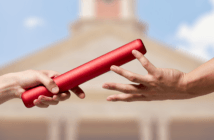 Two people's hands passing a baton from one to the other in front of a blurred image of a church