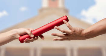 Two people's hands passing a baton from one to the other in front of a blurred image of a church