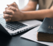 Person with hands clenched in prayer over a laptop keyboard atop a table next to a Bible