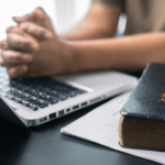 Person with hands clenched in prayer over a laptop keyboard atop a table next to a Bible