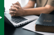 Person with hands clenched in prayer over a laptop keyboard atop a table next to a Bible