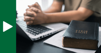 Person with hands clenched in prayer over a laptop keyboard atop a table next to a Bible