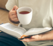 Person sitting on a couch reading a book while drinking a cup of hot tea