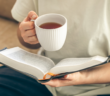 Person sitting on a sofa reading a book while holding a cup of tea