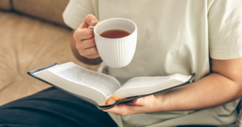 Person sitting on a sofa reading a book while holding a cup of tea